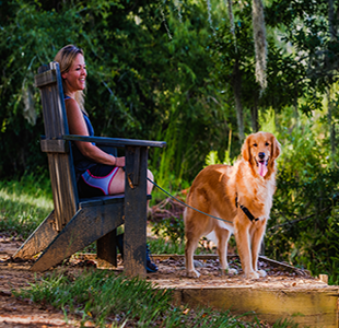 A smiling woman sits in a wooden chair next to her leashed golden retriever at Lafayette Heritage Trail Park.