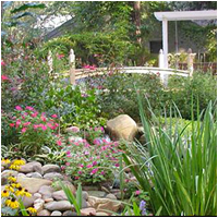 A charming white footbridge crosses a small, rocky stream and is surrounded by lush gardens with pink and yellow flowers at Dorothy B. Oven Park.