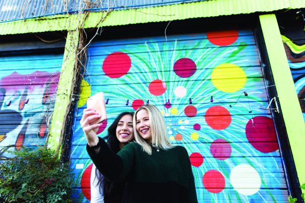 two women take a selfie in front of murals at railroad square art park